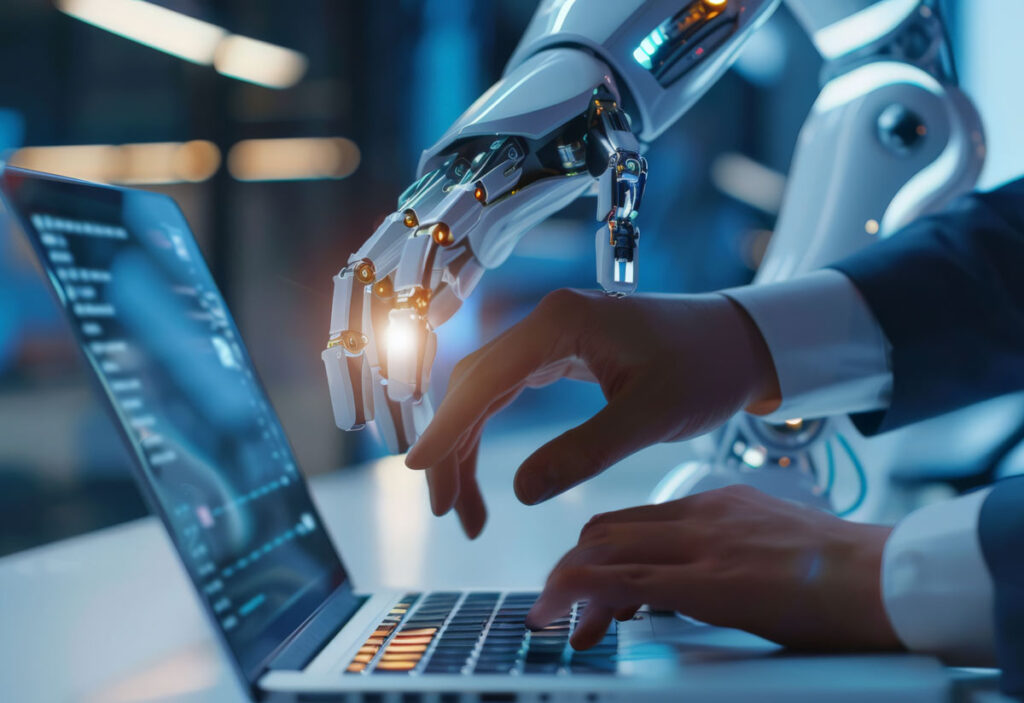 Zdjęcie przedstawia dłonie ludzkie na klawiaturze laptopa ], oraz dłoń robota pokazującą coś na monitorze laptopa.
